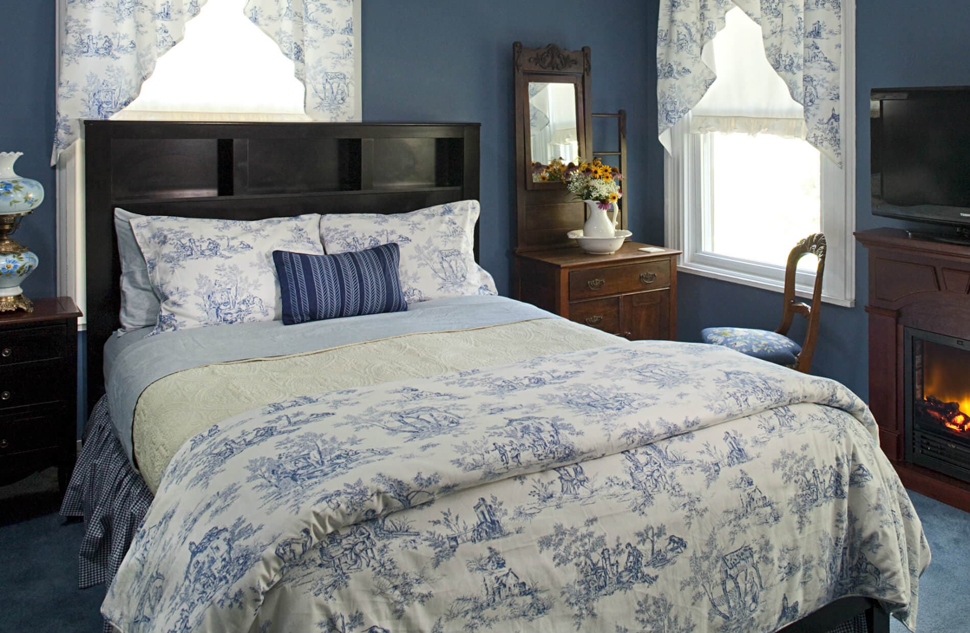 A cozy bedroom features a queen bed with blue and white floral bedding, a dark headboard, a lit fireplace, and an antique dresser.