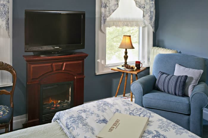 A cozy room with blue walls, a bed with floral bedding, a TV over a lit fireplace, and a blue armchair by a window with a lamp.