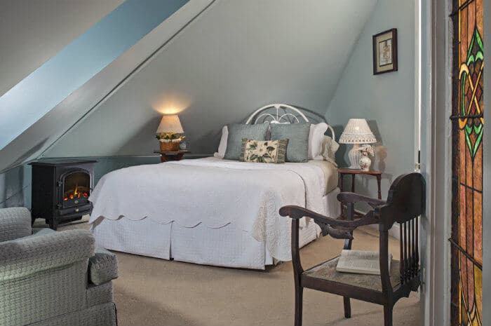 A serene attic bedroom features a white queen bed with light blue accents, a small fireplace, and a stained-glass window, creating a cozy ambiance.