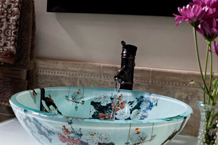 A glass vessel sink with an oriental floral design and a dark, ornate faucet, with purple flowers in a vase beside it.