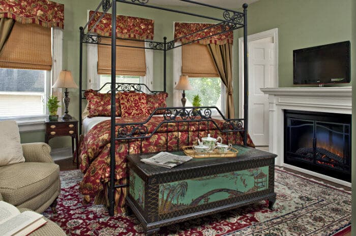 A charming bedroom with a four-poster bed adorned with red and gold bedding. It features a fireplace, TV, and decorative trunk, creating a cozy ambiance.