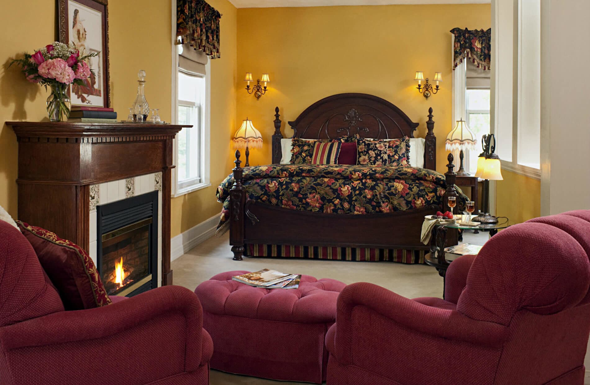 Kind bed with high dark wood headboard; fireplace to left, two chairs