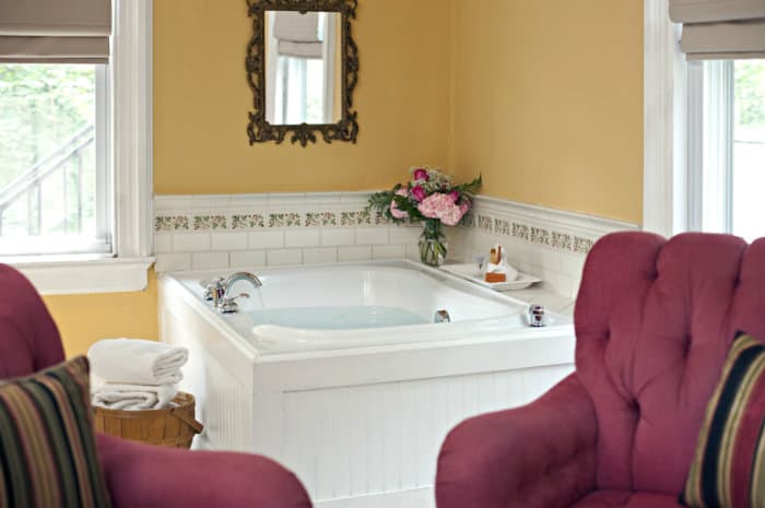 A luxurious corner jetted tub with a bath caddy, flowers, and candles sits in a yellow room between two windows. Two red armchairs are in the foreground.