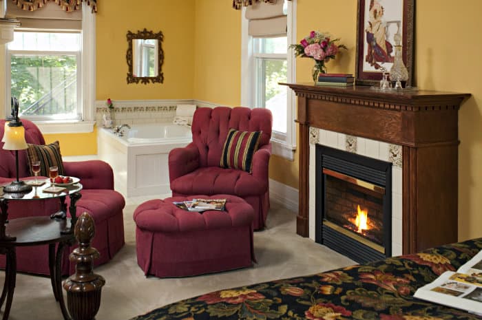 A rich yellow room with a bed, a lit fireplace, two red armchairs and an ottoman, and a Jacuzzi tub by windows.