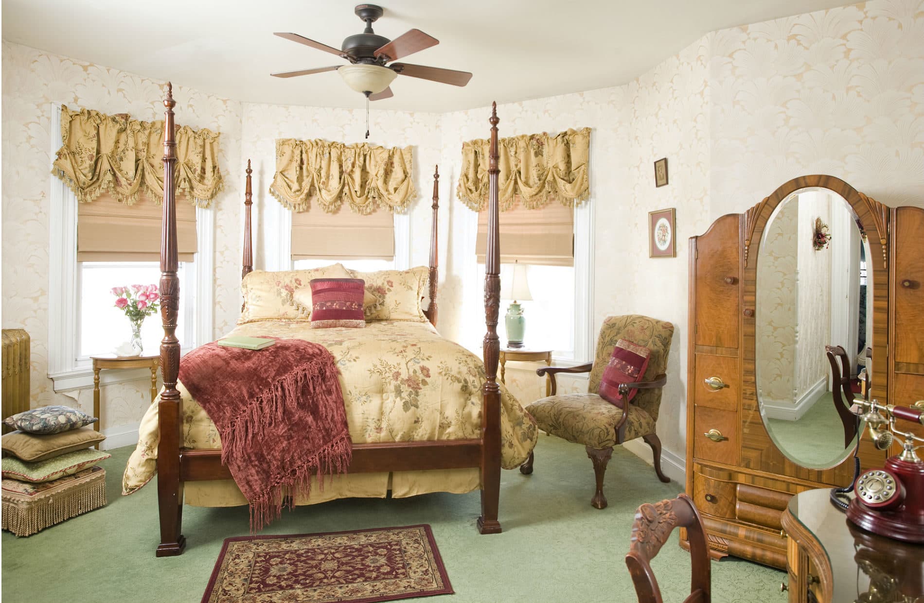 A vintage-style bedroom with a four-poster queen bed, gold and red bedding, an antique dresser with a mirror, and a ceiling fan.