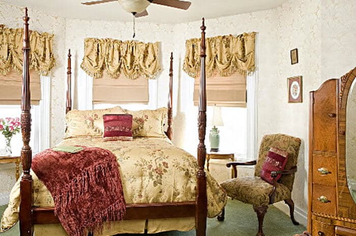 A classic bedroom features a four-poster queen bed with gold and red bedding, an antique dresser, a ceiling fan, and warm lighting.