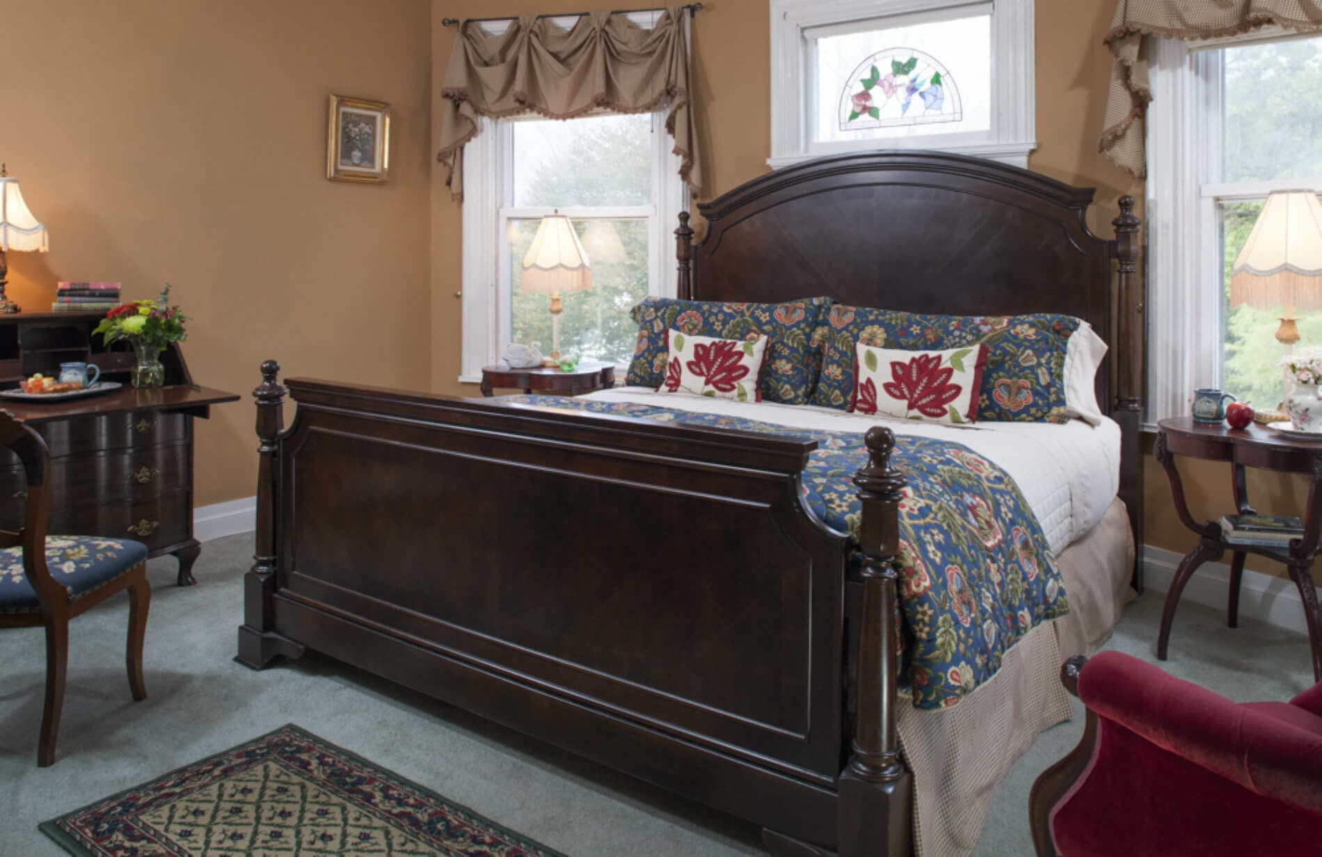 A warm, inviting bedroom with a dark wood king bed featuring blue and red bedding. It includes a desk, a small table, and a stained-glass window.