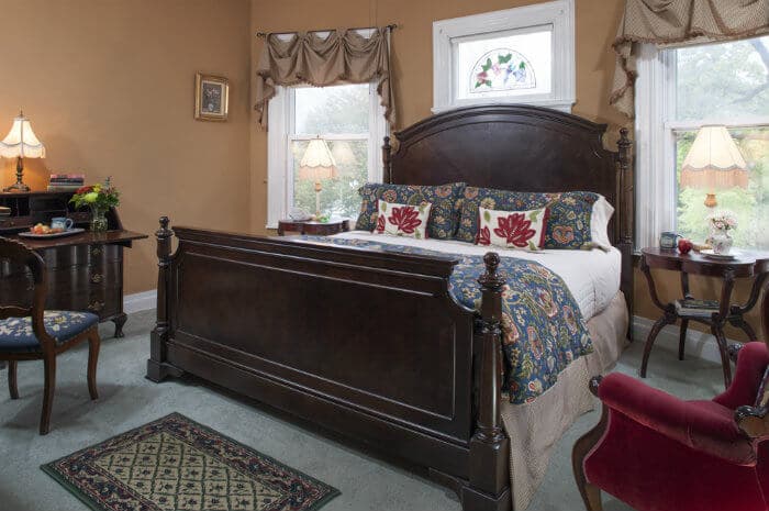 A spacious bedroom with warm taupe walls features a large dark wood king bed with red & blue floral bedding, surrounded by elegant furniture and two bright windows.