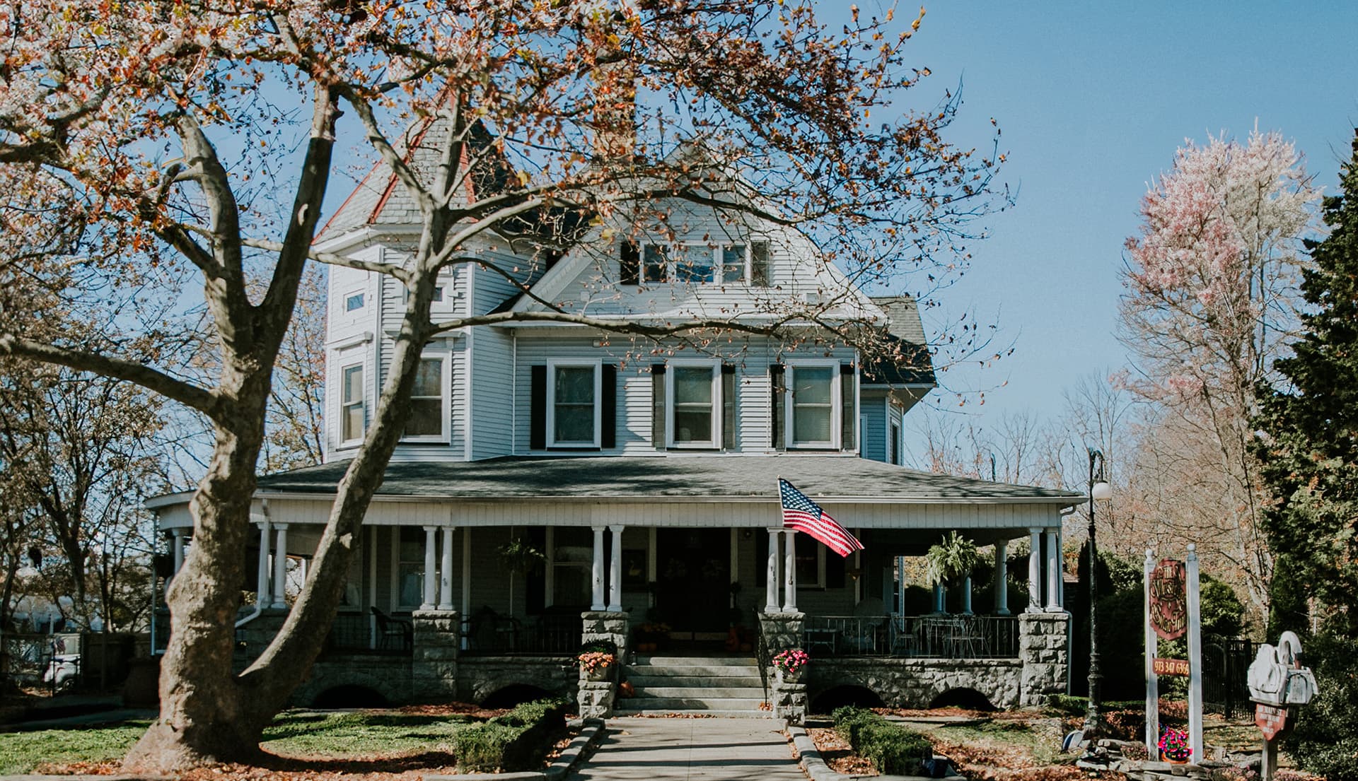 A multi-story Victorian house with a front porch, an American flag, and colorful flowers in autumn.