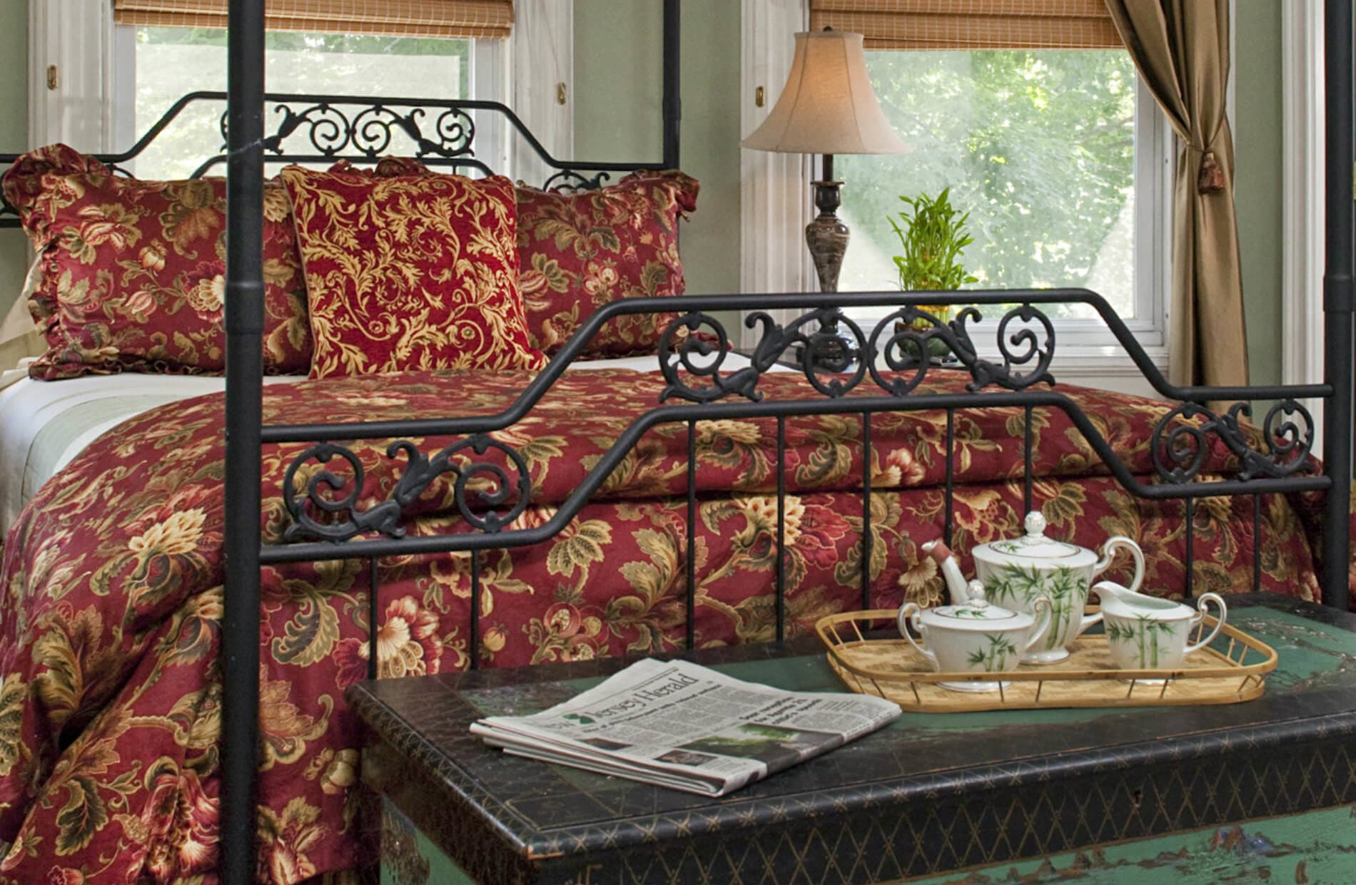 A regal bedroom with a black wrought-iron four-poster queen bed, adorned with rich red and gold patterned bedding. A tea set sits on an oriental trunk in front of the bed.