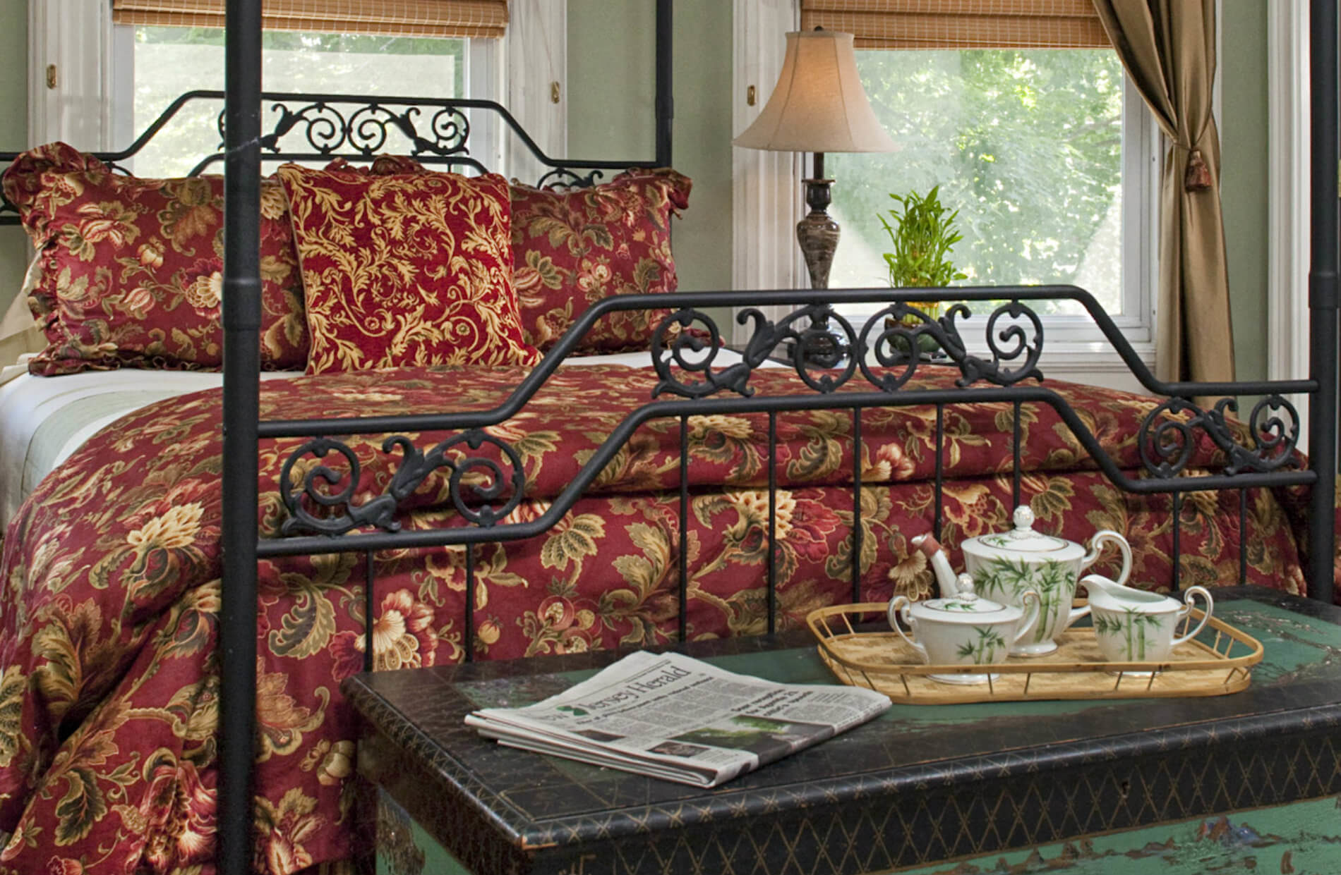 A regal bedroom with a black wrought-iron four-poster queen bed, adorned with rich red and gold patterned bedding. A tea set sits on an oriental trunk in front of the bed.