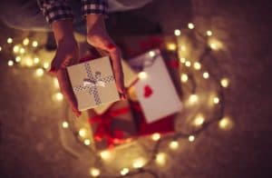 A person holds a gift box surrounded by wrapped presents and glowing lights.