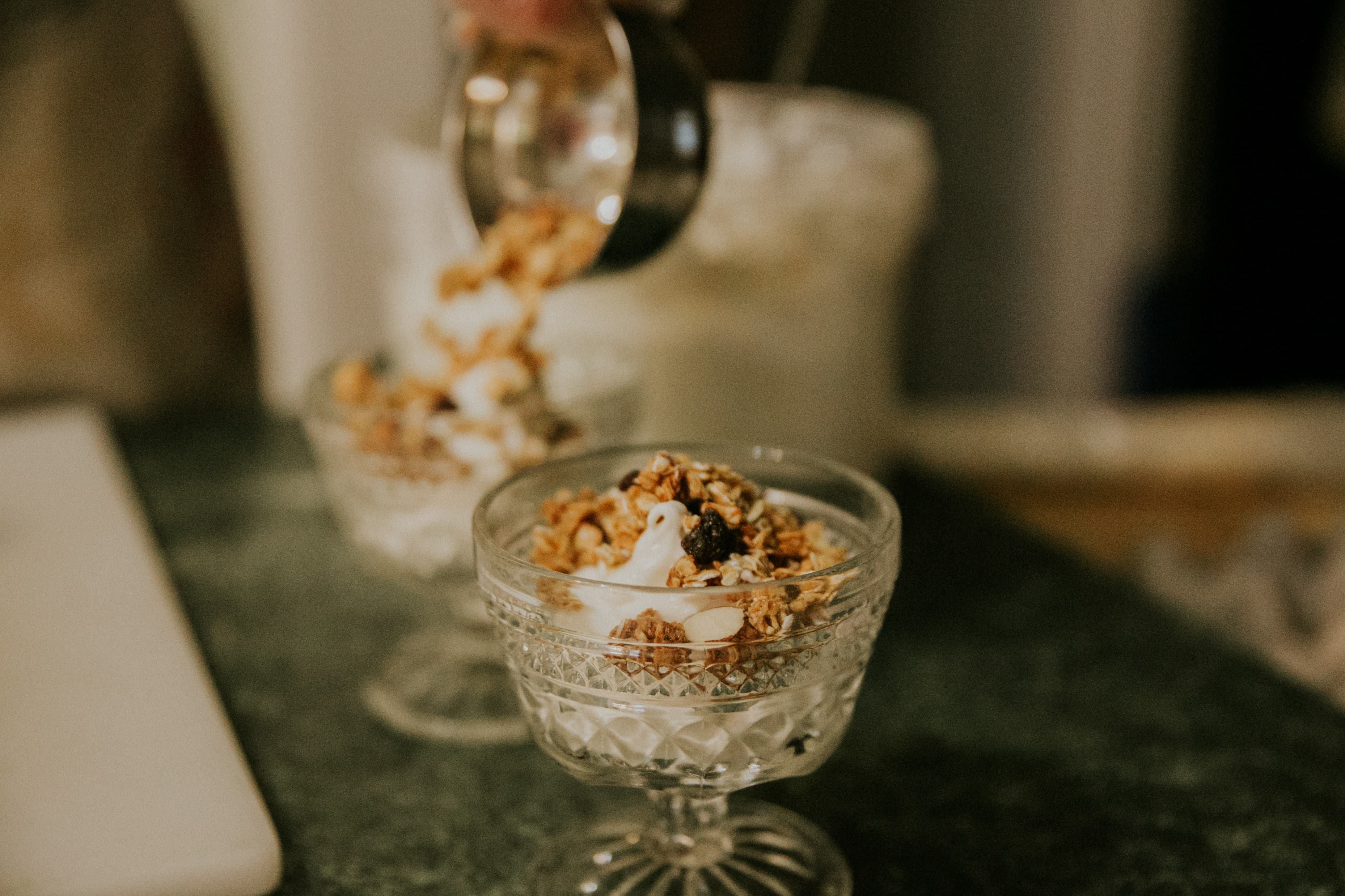 A hand is pouring granola into a glass dessert cup filled with yogurt.