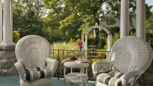 Two white wicker chairs and a small table on a porch overlooking a lush garden.