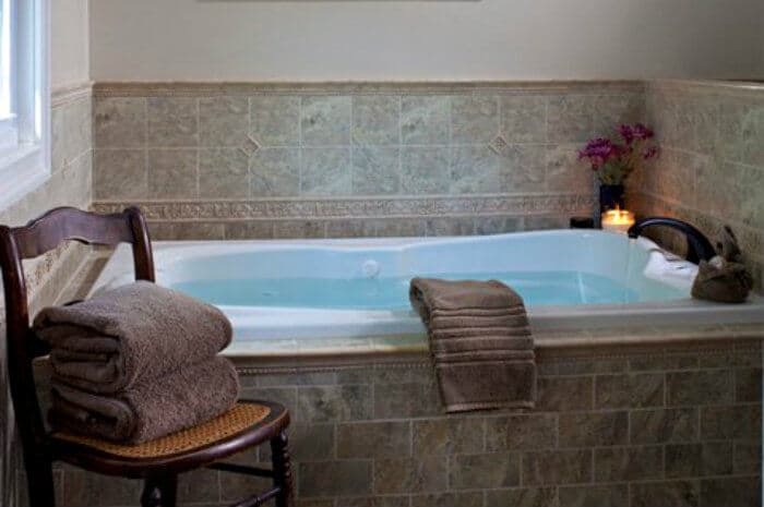 A tranquil bathroom with a large, tiled two-person Jetted tub filled with water, next to a wooden chair holding neatly folded brown towels.