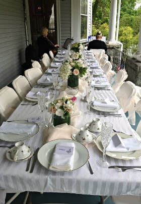 A elegantly set long table with floral centerpieces, fine china, and crystal glasses on a porch.