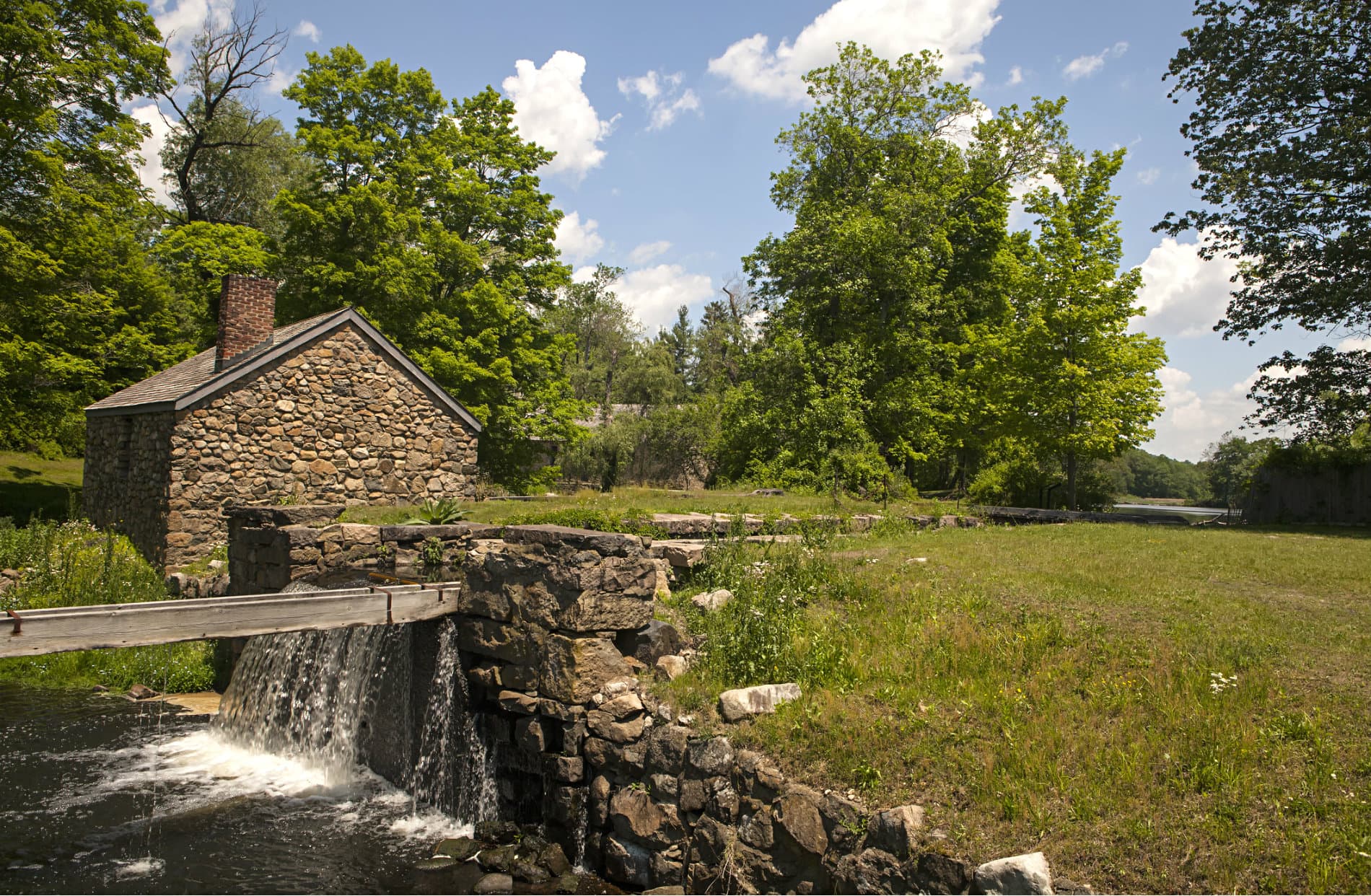A stone building near a waterfall and lush green trees under a blue sky.