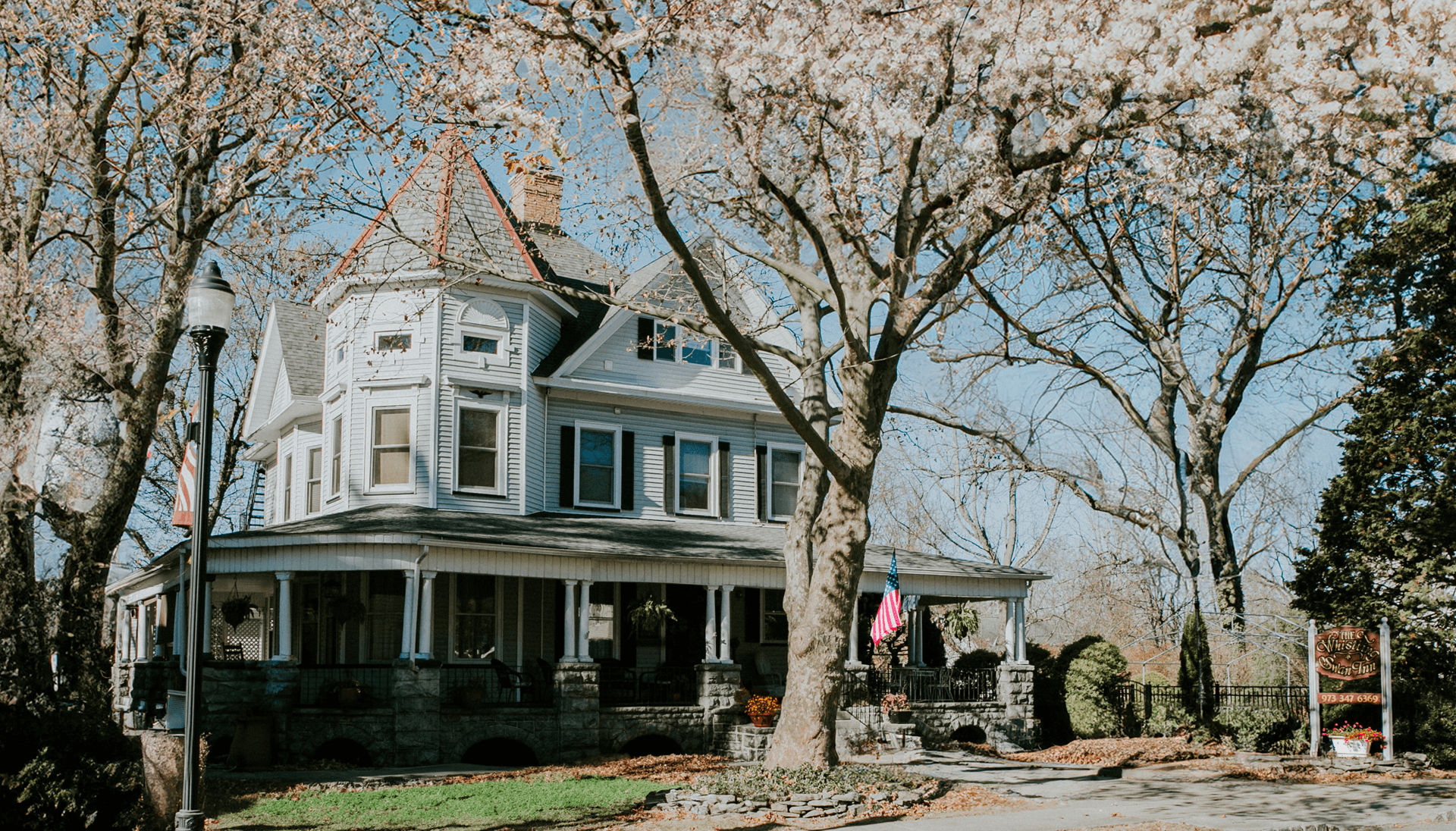 A multi-story Victorian house with a front porch, an American flag, and colorful flowering trees in spring.