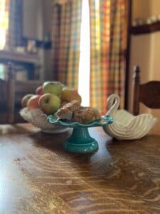 A table set with a turquoise pedestal dish of cookies and a bowl of fresh apples, with colorful curtains in the background.