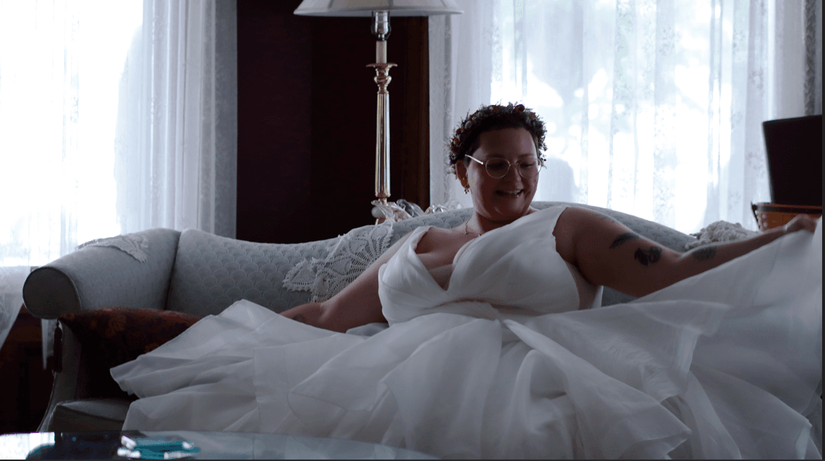 A smiling person in a white wedding dress sits on a couch, adjusting their gown.