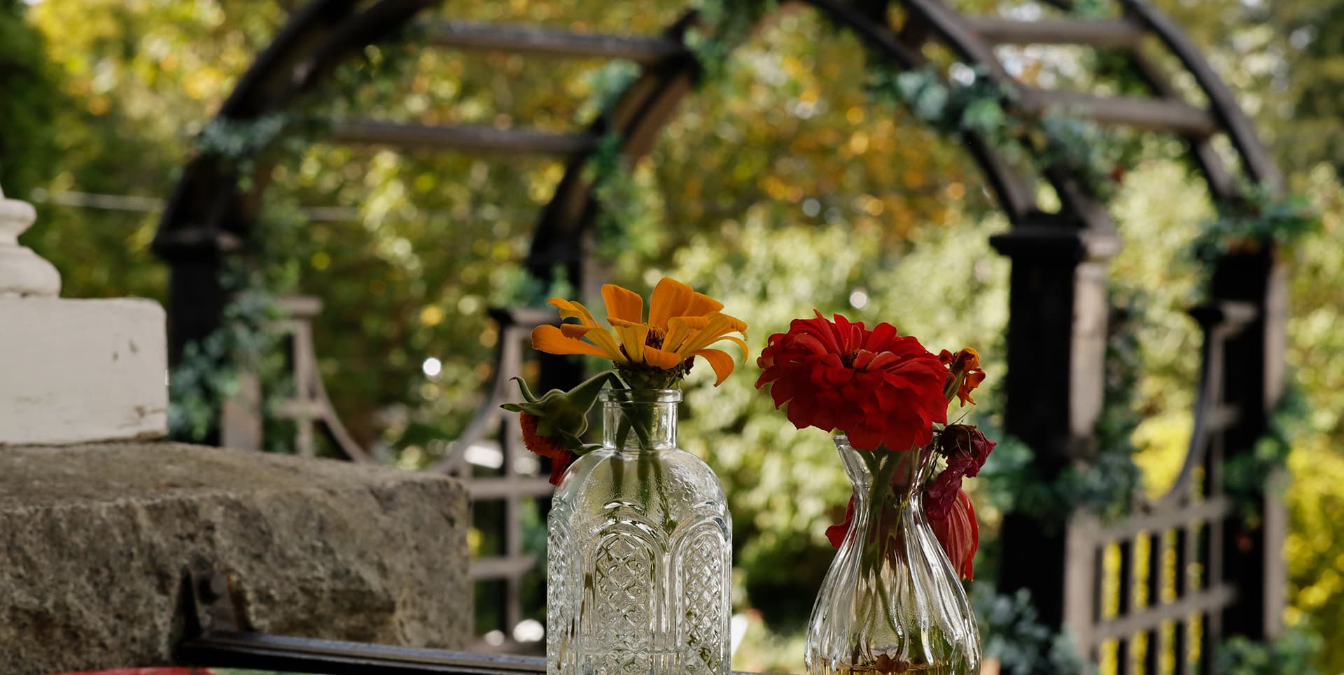 Two vases with colorful flowers are placed on a stone surface, with a garden arbor in the background.