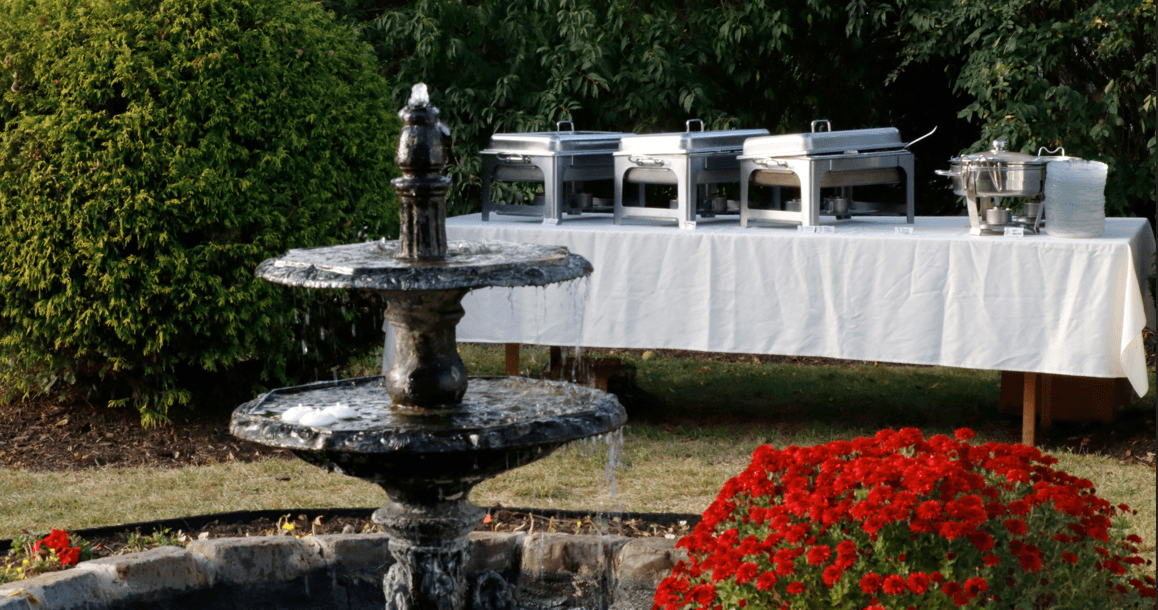 A decorative fountain in the foreground with a buffet table and red flowers in a garden setting.