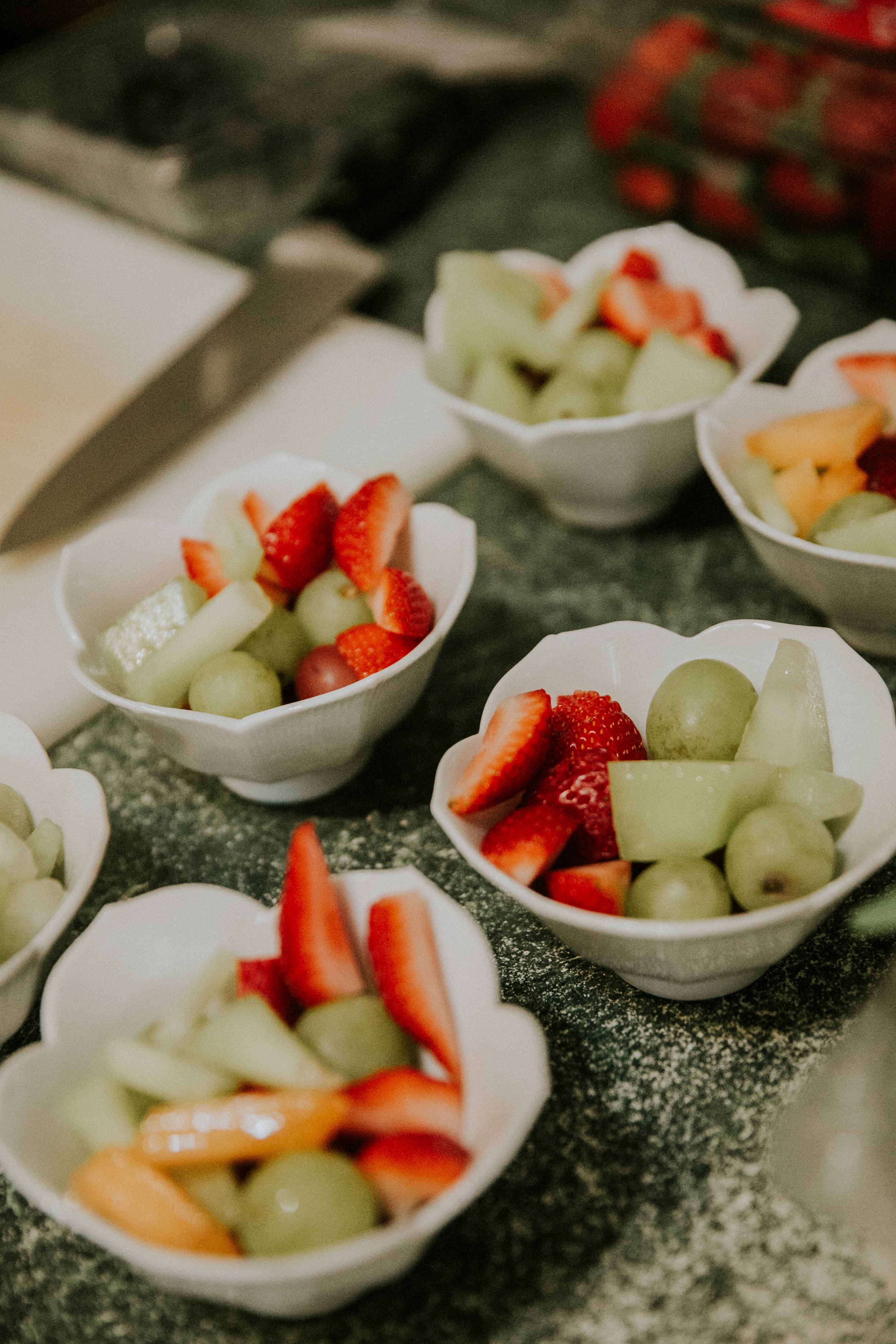 Several small white bowls filled with assorted fruit, including strawberries, melon, and grapes, arranged on a countertop.