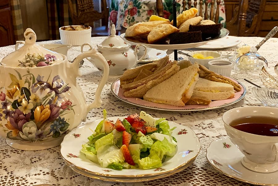 A beautifully arranged tea table featuring a floral teapot, assorted sandwiches, scones, a fresh salad with strawberries, and a cup of tea.
