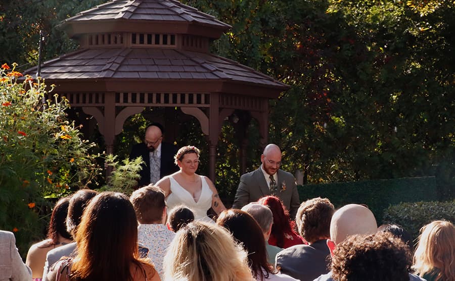 A couple walks down the aisle in front of a gazebo during a wedding ceremony, with guests seated in the foreground.
