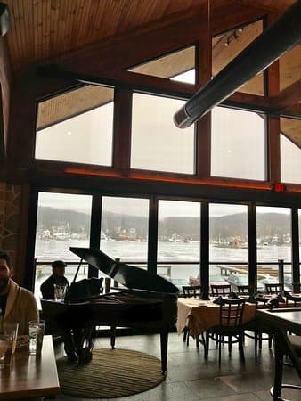 A cozy restaurant interior featuring a grand piano, large windows overlooking a lake, and diners enjoying their meals.