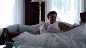 A smiling person in a white wedding dress relaxes on a sofa in a bright room.