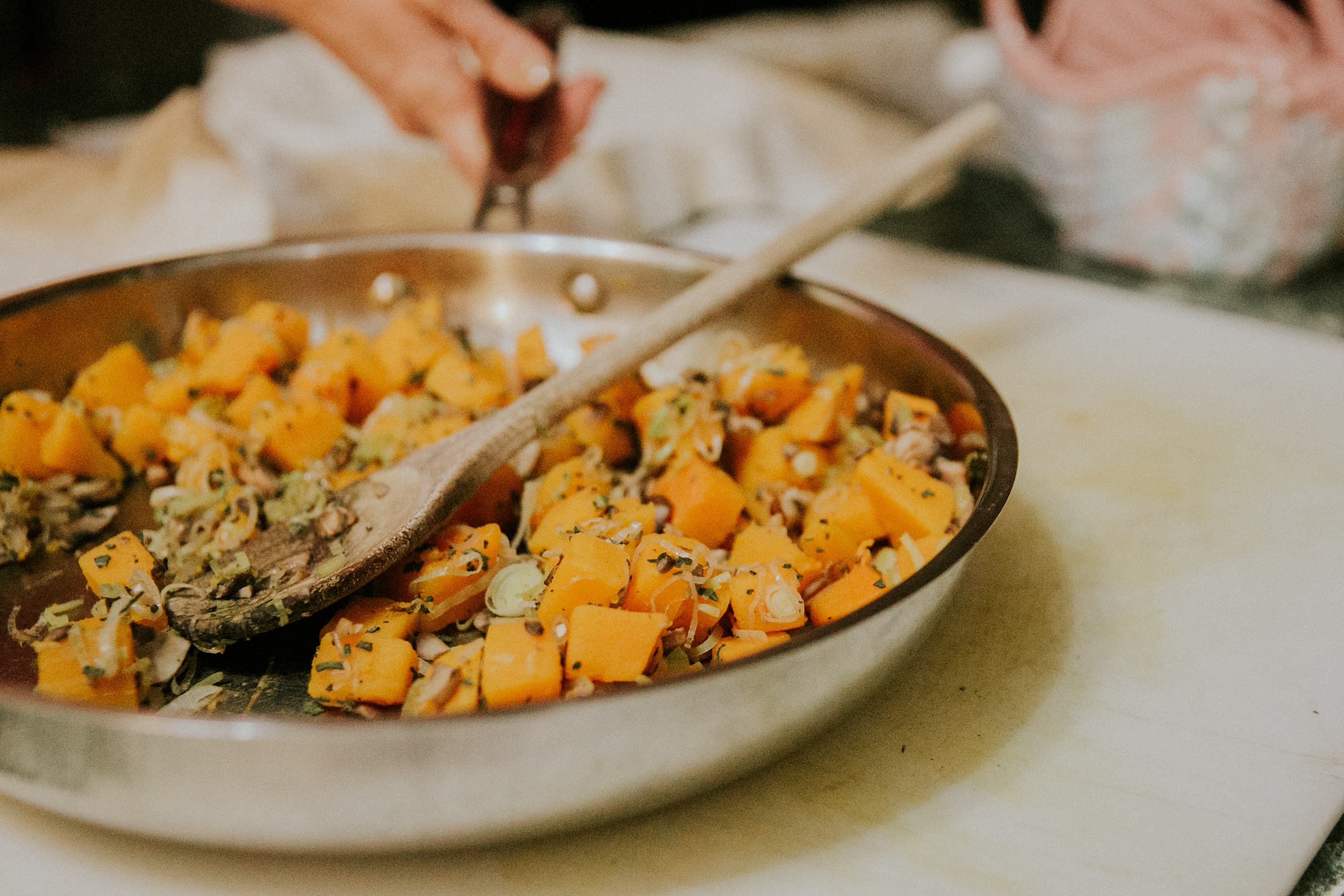 A hand stirs sautéed butternut squash and vegetables in a pan with a wooden spoon.