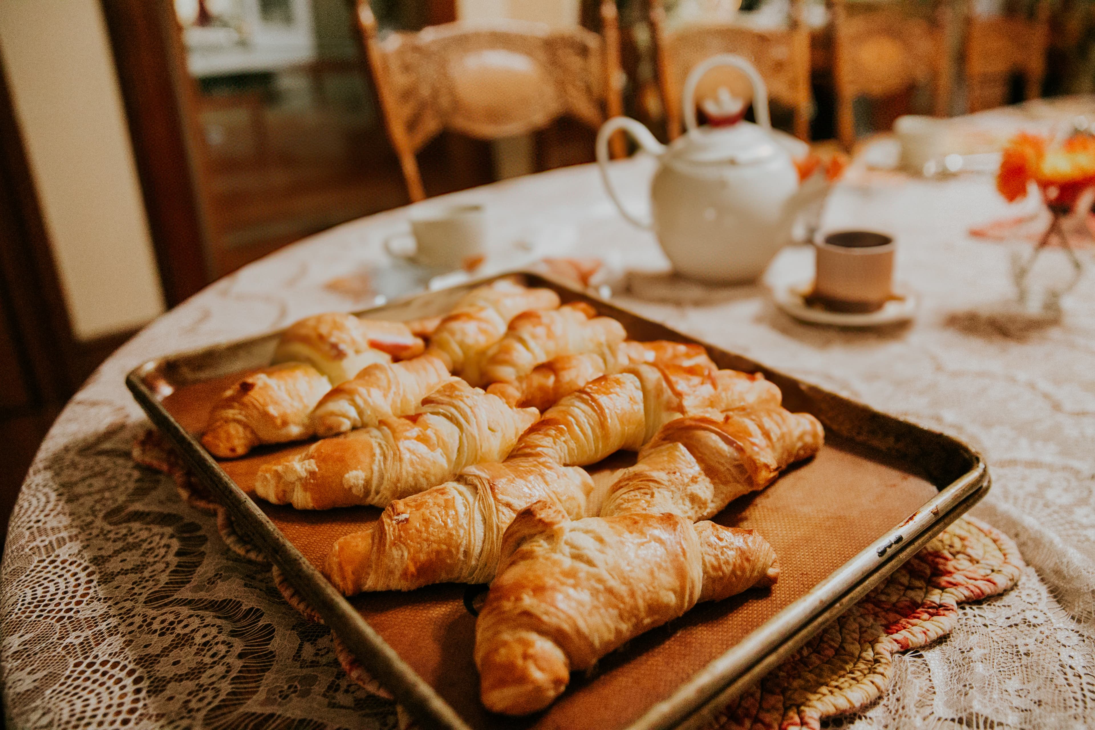 A tray of freshly baked croissants sits on a lace-covered table with a teapot and cup in the background.