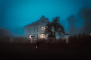 A foggy night scene featuring a large, illuminated house surrounded by trees.
