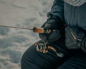 A person in gloves holds a fishing rod and reel while sitting on snow.