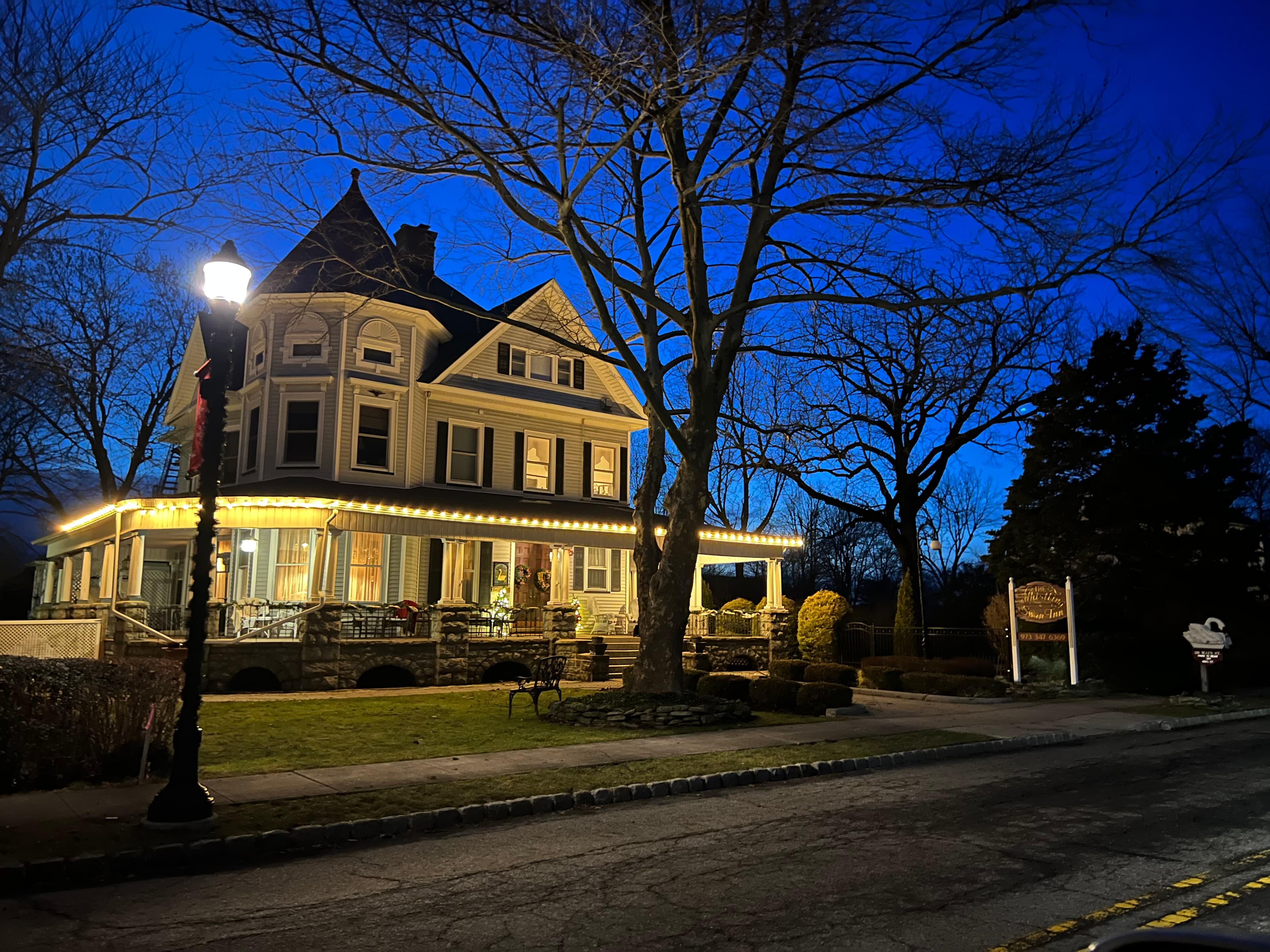 A large, beautifully lit Victorian house stands beside a quiet street at dusk, surrounded by trees.