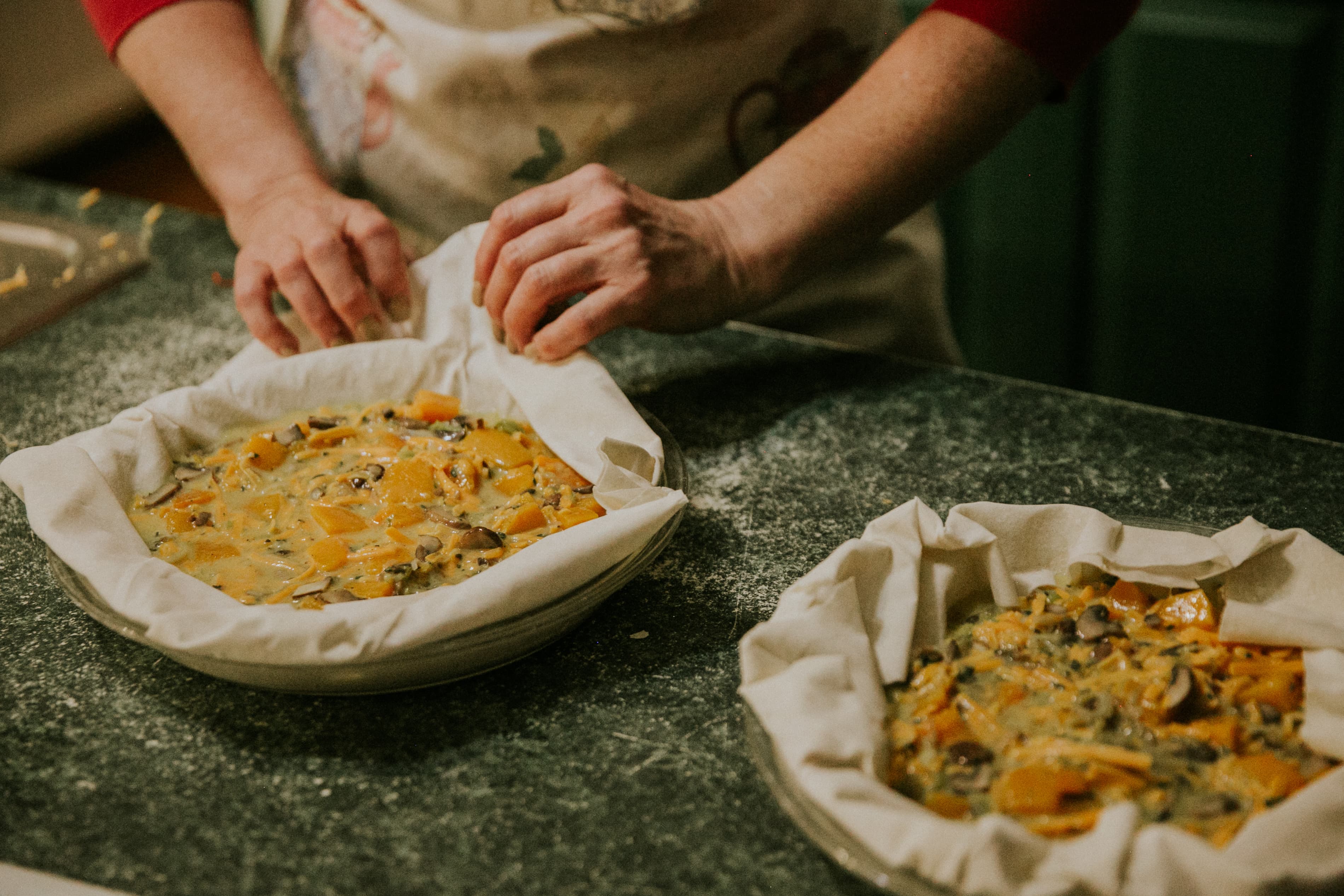 A person prepares two pie dishes lined with dough and filled with colorful ingredients on a floured countertop.