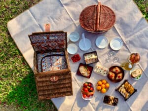 A picnic spread featuring a wicker basket, plates, and various delicious foods on a blanket in the grass.