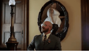 A man in a suit smiles in front of a mirror reflecting a woman in a white dress.