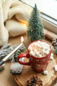 A festive red mug filled with marshmallows sits on a wooden surface surrounded by holiday decorations and a lit match.