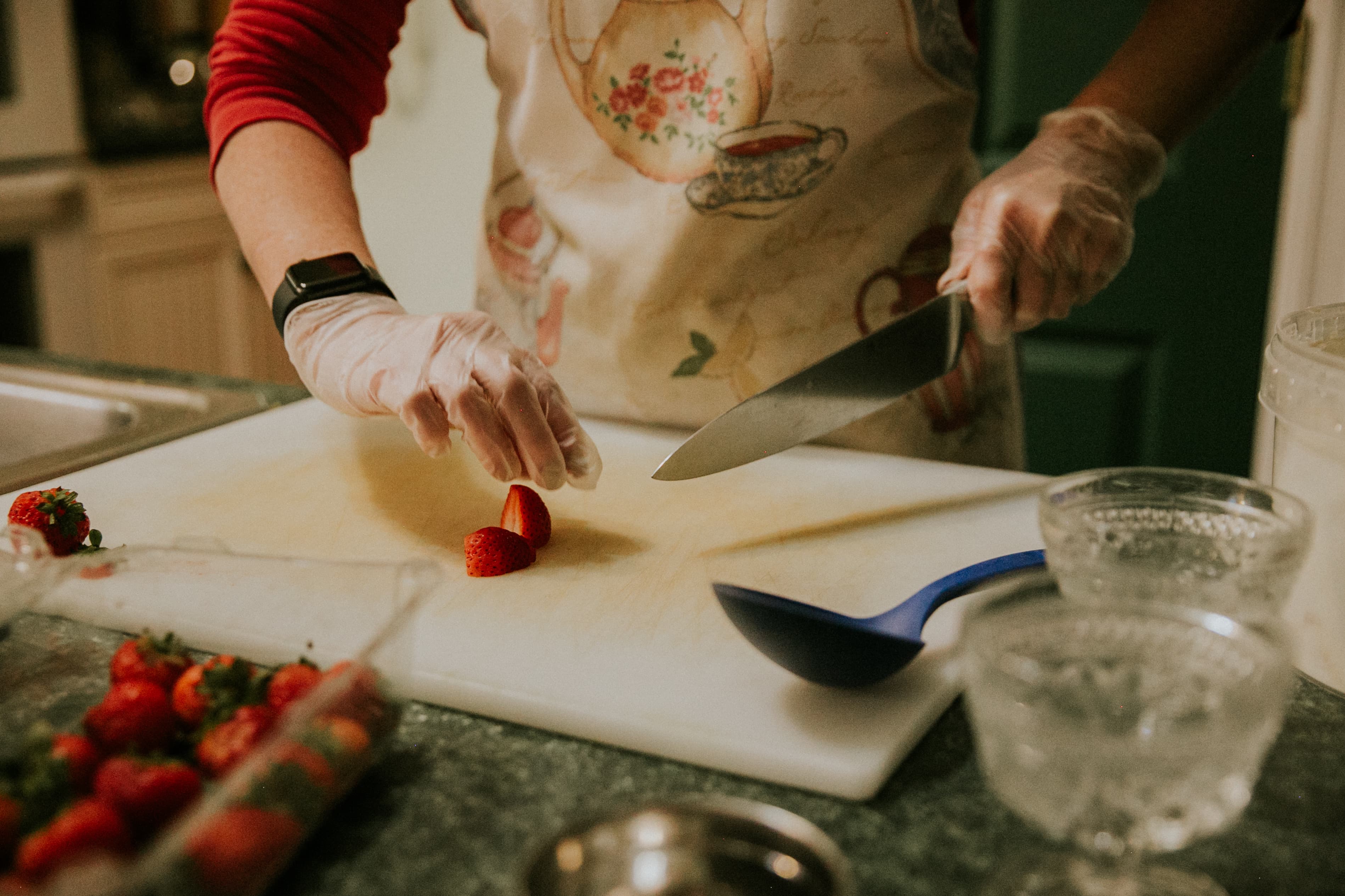 A hand in a glove slices strawberries on a cutting board in a kitchen.