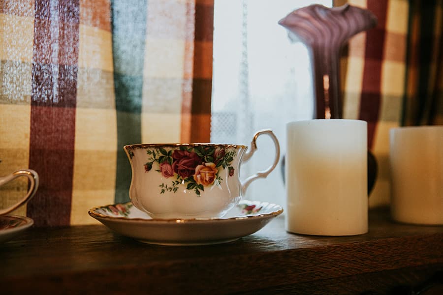 A floral-patterned teacup and saucer sit on a wooden shelf beside candles, with plaid curtains in the background.