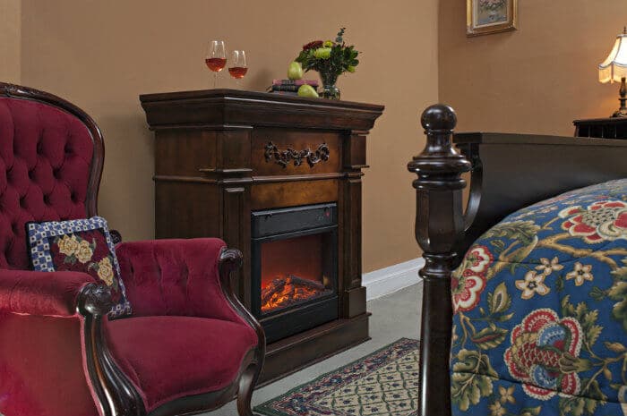 A cozy corner of a bedroom features a lit fireplace, a red armchair, and a small table with wine glasses and fruit, creating a warm ambiance.