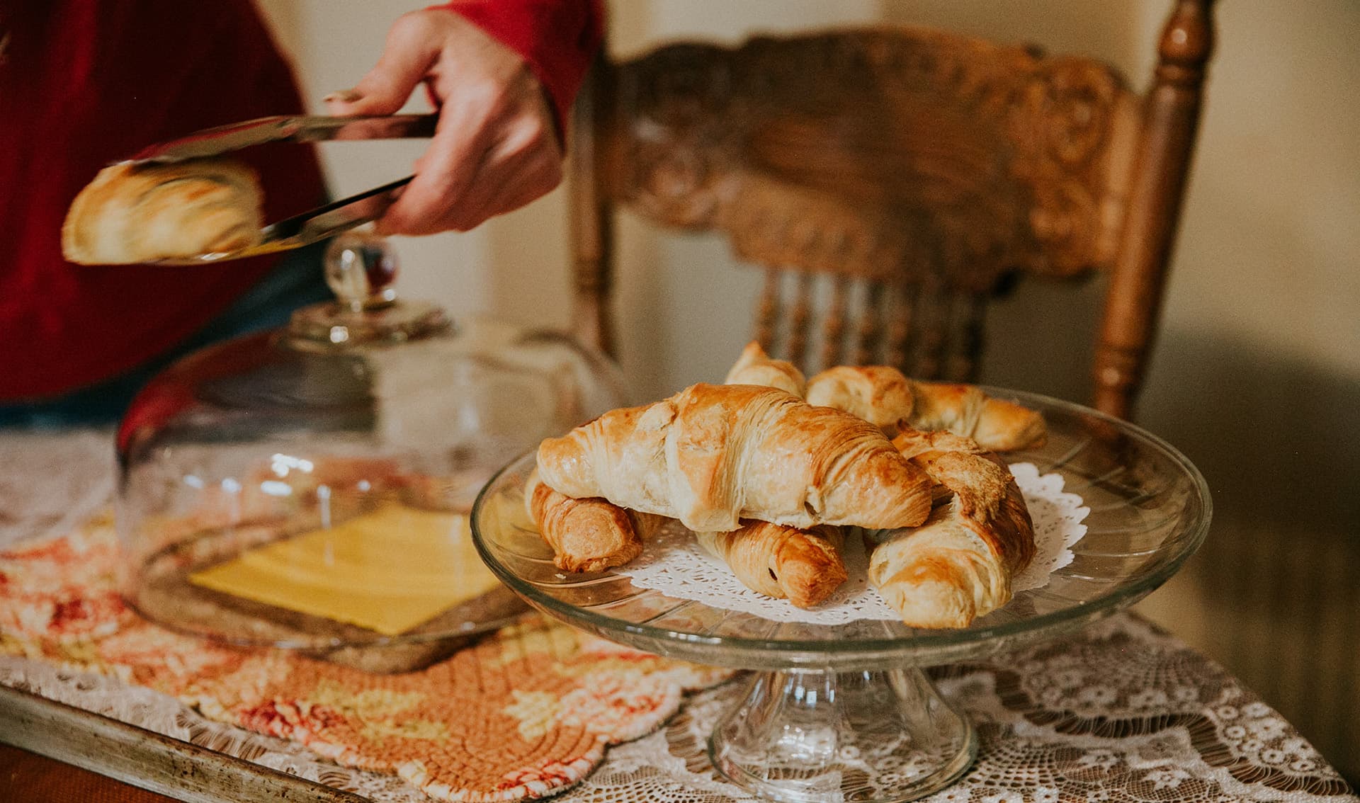 A hand uses tongs to serve freshly baked croissants from a glass cake stand.