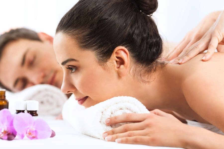 A woman receives a massage while a man relaxes nearby, surrounded by essential oils and a flower.