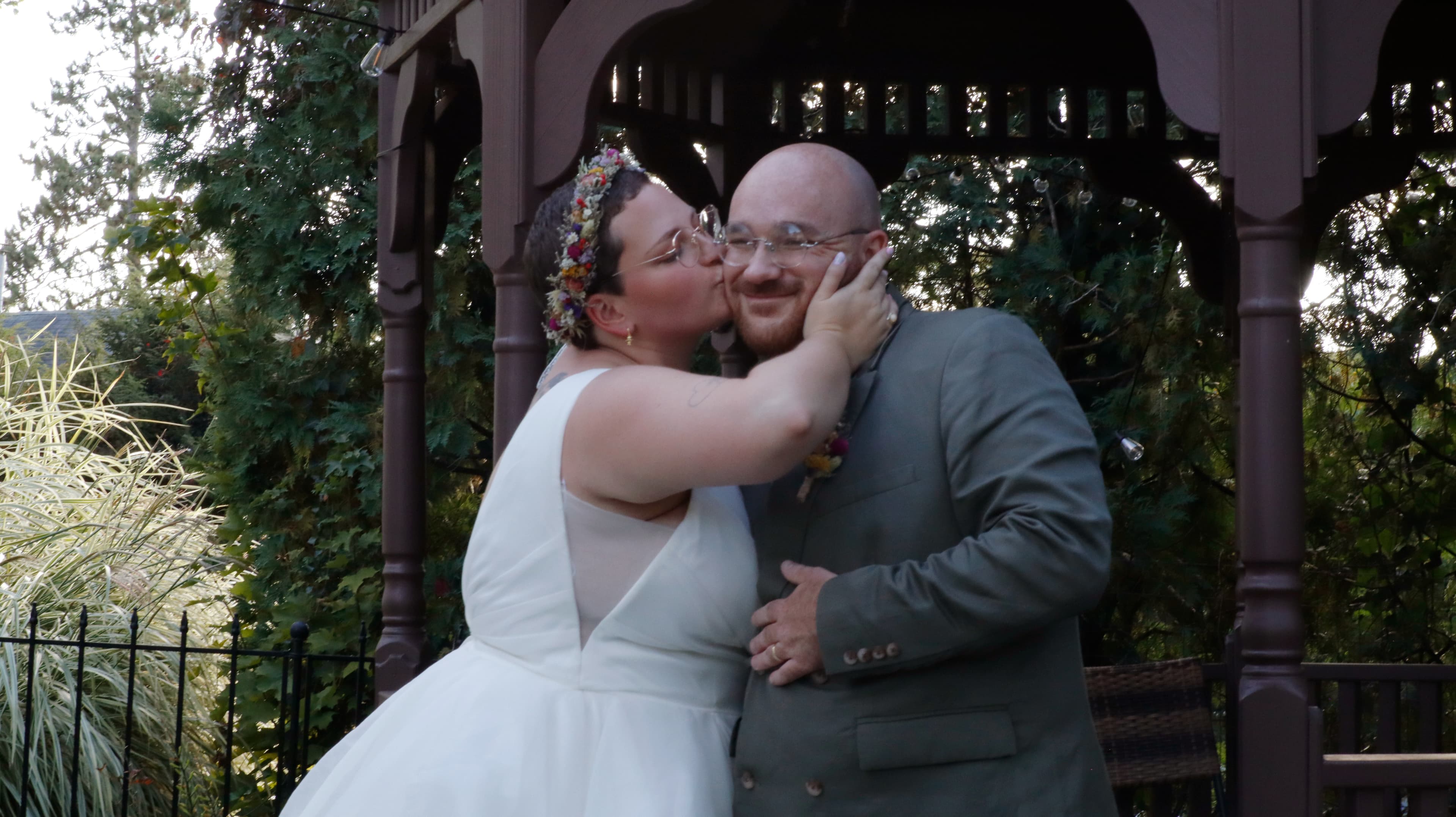 A bride kisses her groom under an outdoor gazebo surrounded by greenery.