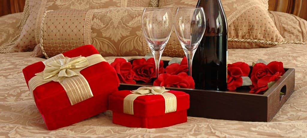 A romantic arrangement featuring two red gift boxes, two wine glasses, a bottle of wine, and scattered rose petals on a decorative platter.