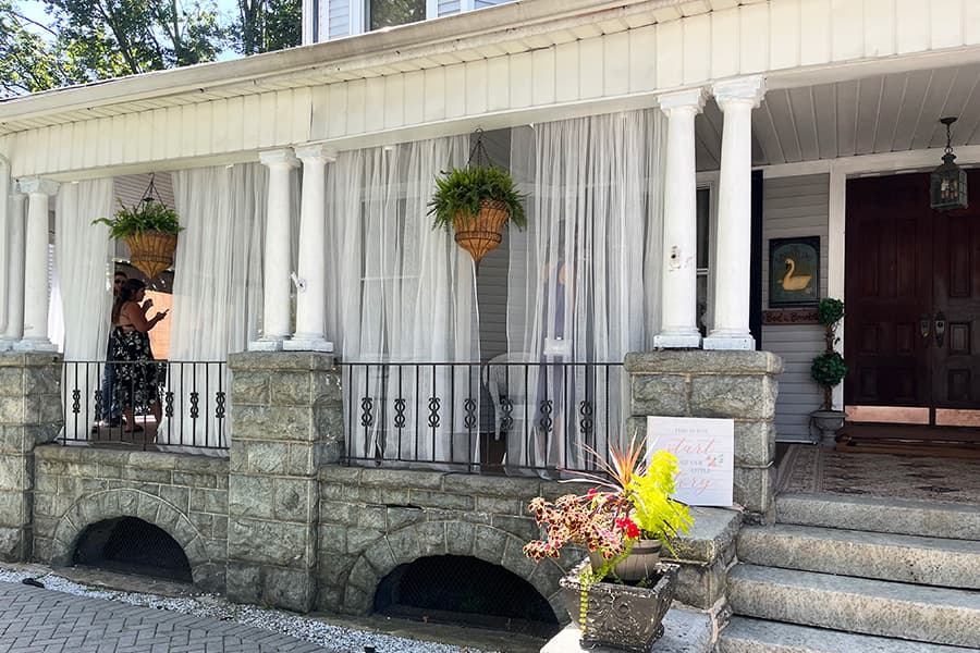 A woman stands on a porch with white curtains and hanging plants beside decorative stone columns.