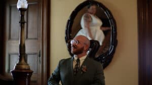 A man in a green suit smiles while standing in front of a mirror reflecting a woman in a white dress.