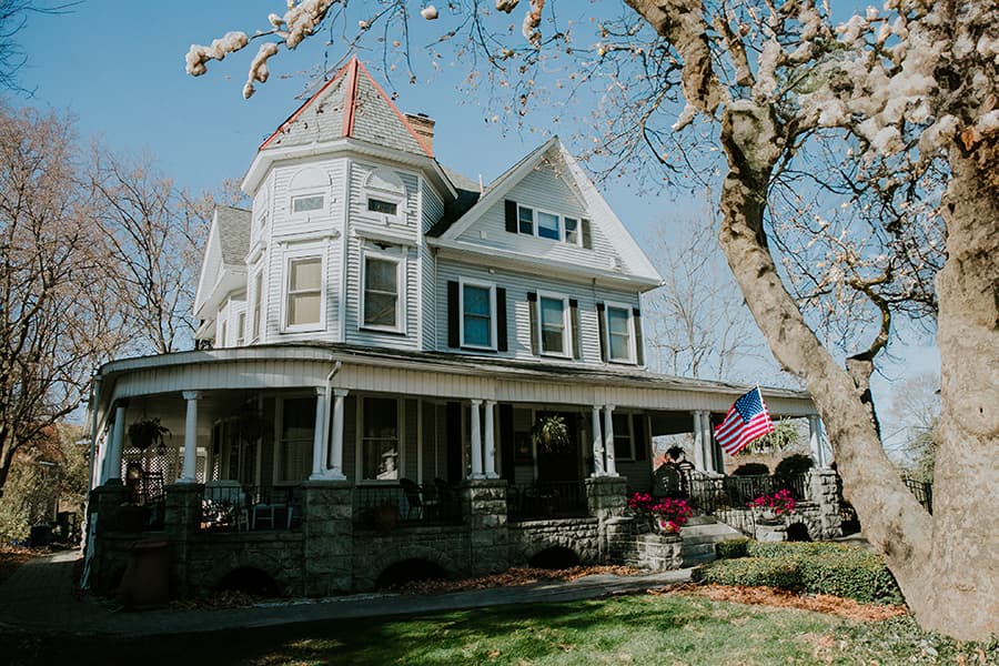 A large Victorian-style house with a front porch, an American flag, and colorful flowers.
